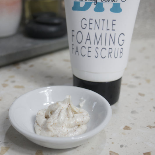 A close up image of a small white dish filled with an off-white product with brown flecks.  Behind is a white tube labelled as Gentle Foaming Face Scrub