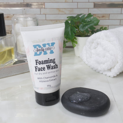 On a bathroom countertop stands a white squeeze tube with a black flip top cap.  The tube has a decorative label identifying it as Foaming Face Wash. Next to the tube is a small black, flat rock that has a puddle of translucent, gel like liquid on it.