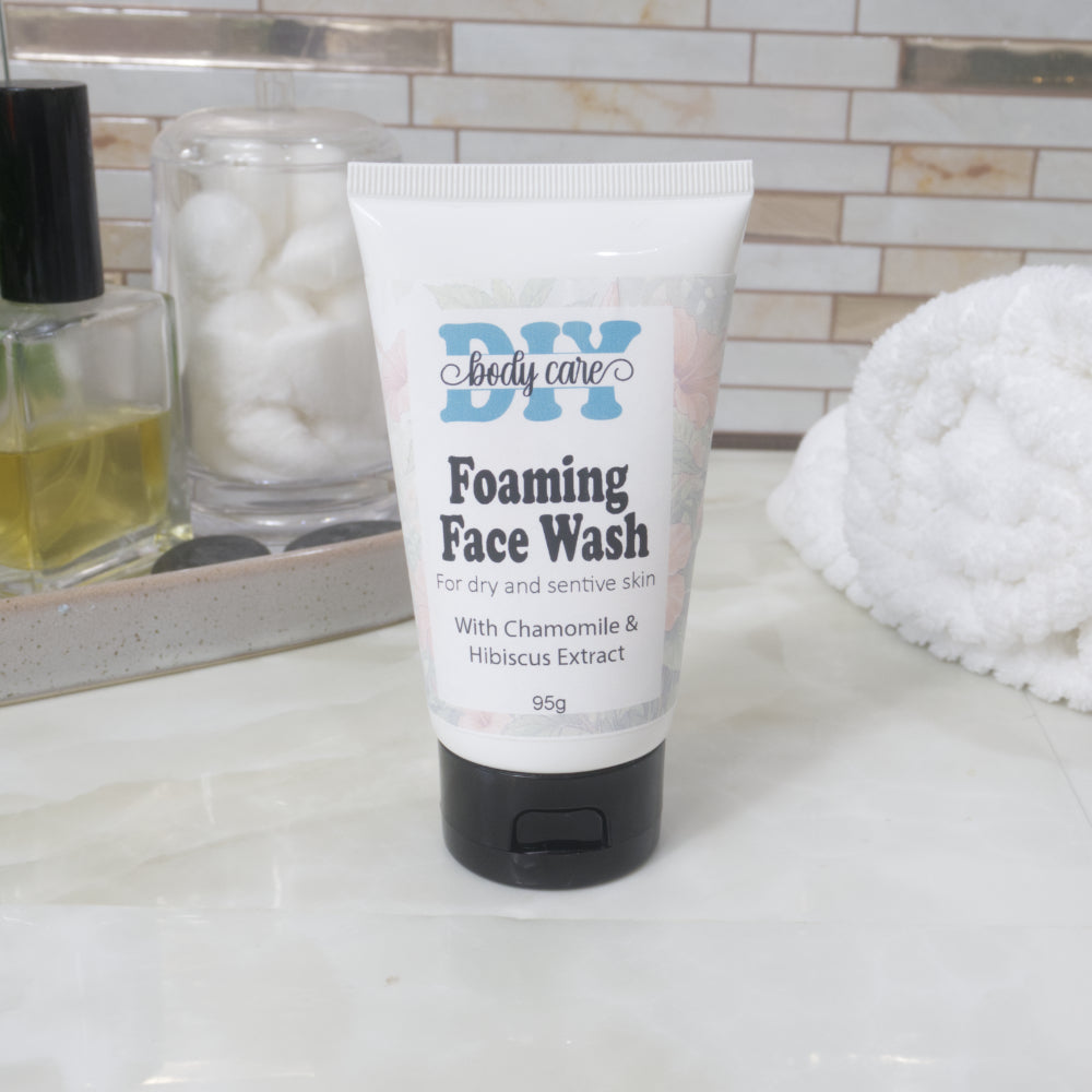 A white squeeze tube with a black cap sits on a bathroom countertop next to a white rolled up towel and a tray with perfume and cotton balls.   The tube has a decorative product label identifying it as Foaming Face Wash