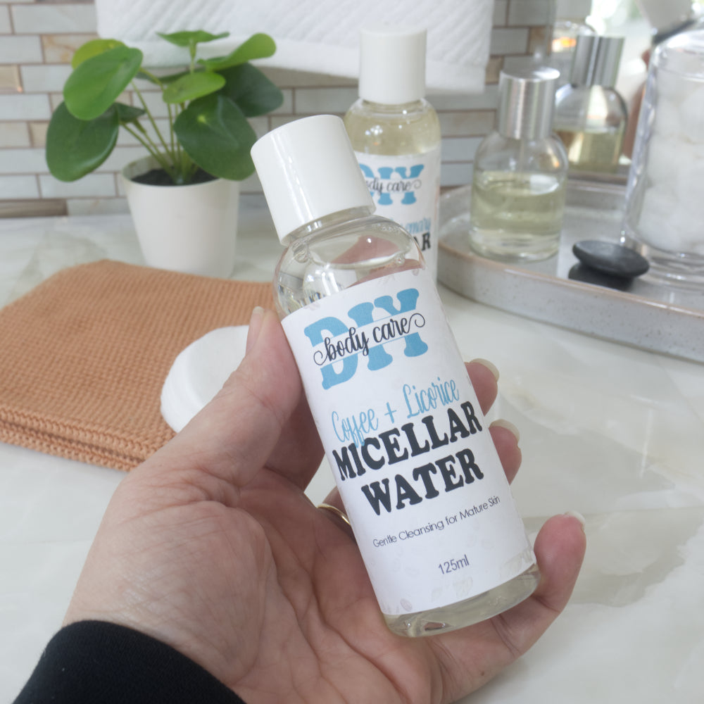 A hand holds a clear bottle with white cap, filled with clear liquid.  It is labelled as Coffee & Licorice Micellar Water.  Behind is a bathroom countertop featuring a face cloth, plant and bottle of perfume.