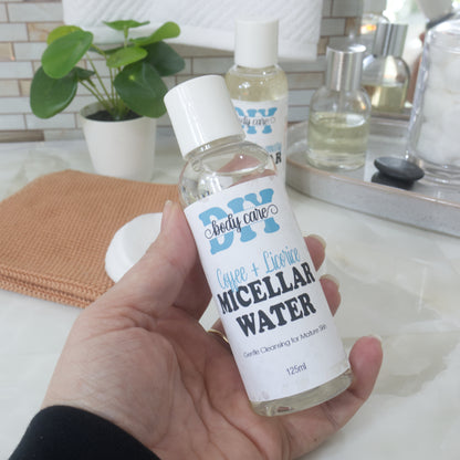 A hand holds a clear bottle with white cap, filled with clear liquid.  It is labelled as Coffee & Licorice Micellar Water.  Behind is a bathroom countertop featuring a face cloth, plant and bottle of perfume.