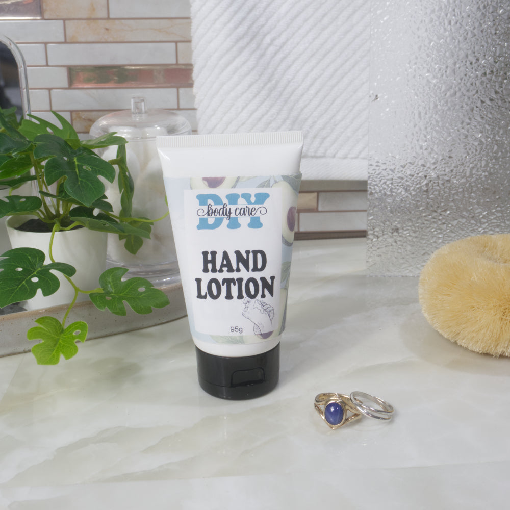 A white tube with black flip top cap stands on a bathroom countertop next to some gold and silver rings.  In the background is a plant, a jar of cotton balls and a towel.  The tube features a decorative product label identifying it as Hand Lotion.