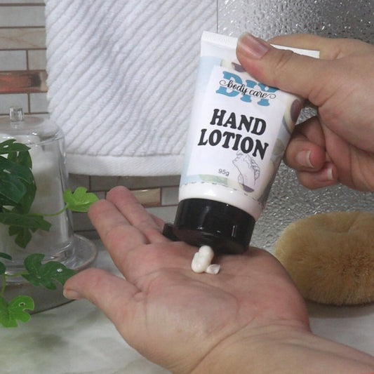 A hand holds a white tube with black flip top cap against the palm of another hand.  A white cream is squeezed out into the palm.  The tube has a product label identifying it as Hand Lotion.  In the backgroun dis a bathroom countertop