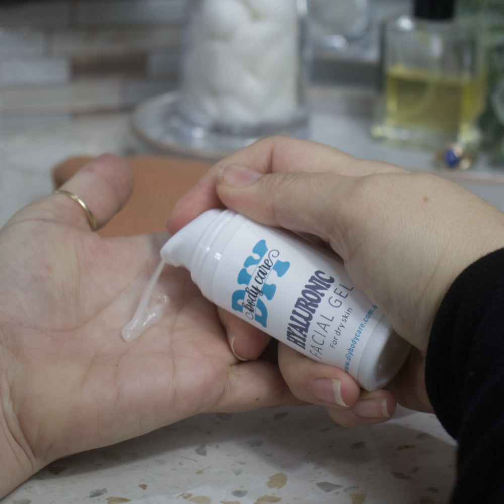 A person is pumping a white bottle of Hyaluronic Facial Gel into the palm of another hand.  The gel has a white, translucent look.  The background is slightly blurred witha bottle of perfume and jar of cotton balls on a stone tray.