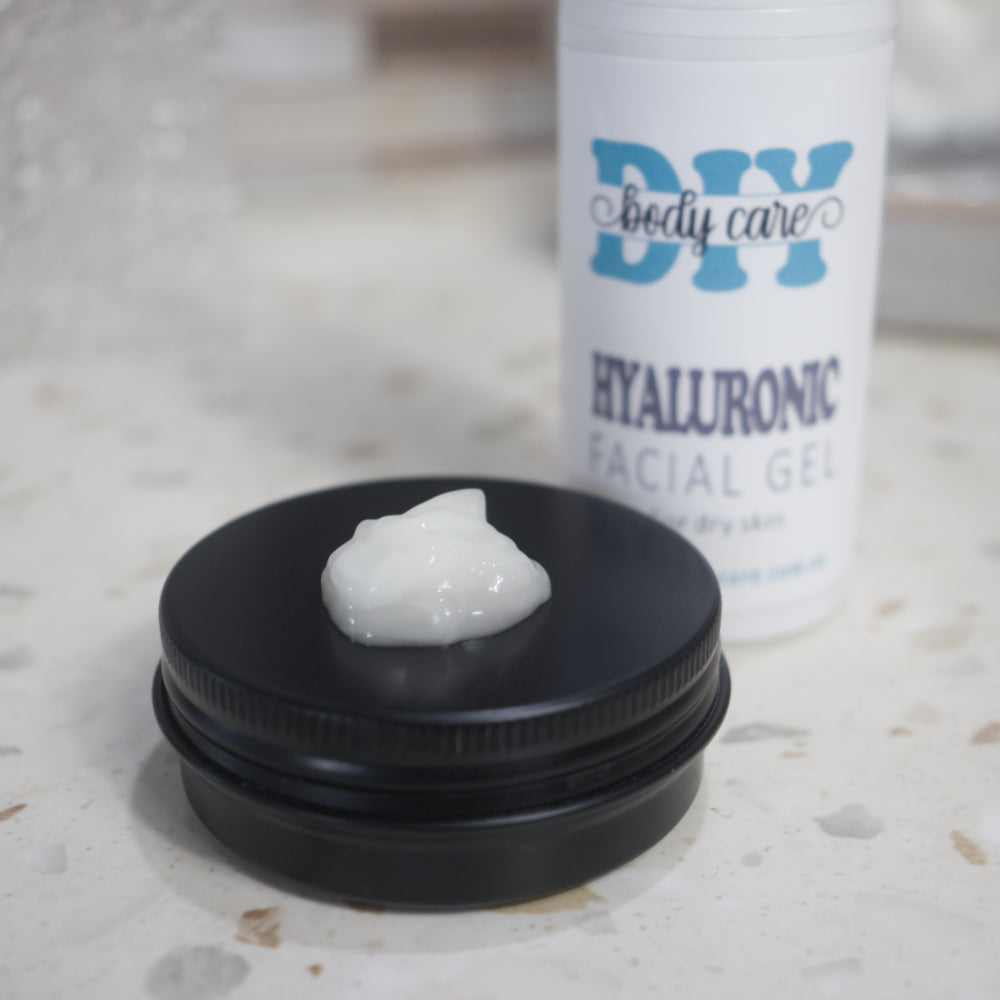 A close up image of a black tin with lid on and a small amount of white gel sitting in the center.  Behind is a white bottle labelled Hyaluronic Facial Gel with the rest of the background heavily blurred.