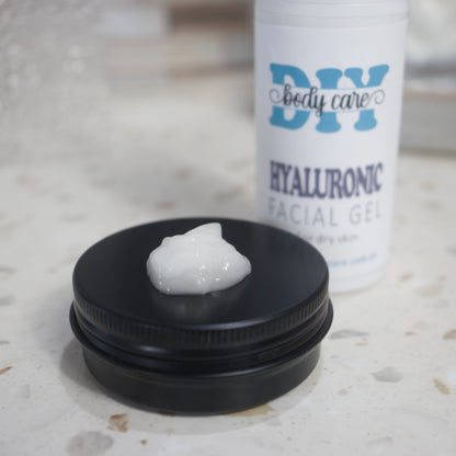 A close up image of a black tin with lid on and a small amount of white gel sitting in the center.  Behind is a white bottle labelled Hyaluronic Facial Gel with the rest of the background heavily blurred.