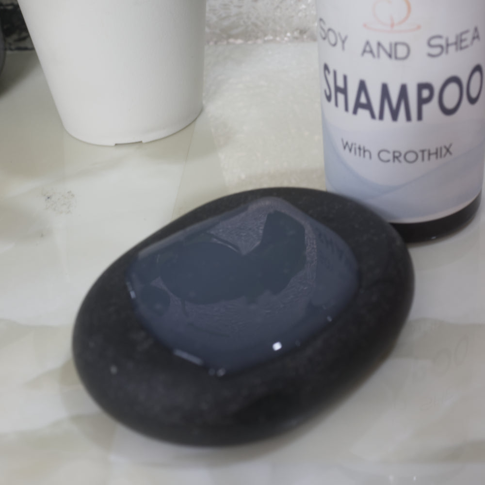 A close up image of a black massage stone with a white translucent liquid on it.  Behind is a bottle labelled SHAMPOO