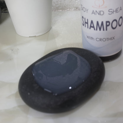 A close up image of a black massage stone with a white translucent liquid on it.  Behind is a bottle labelled SHAMPOO