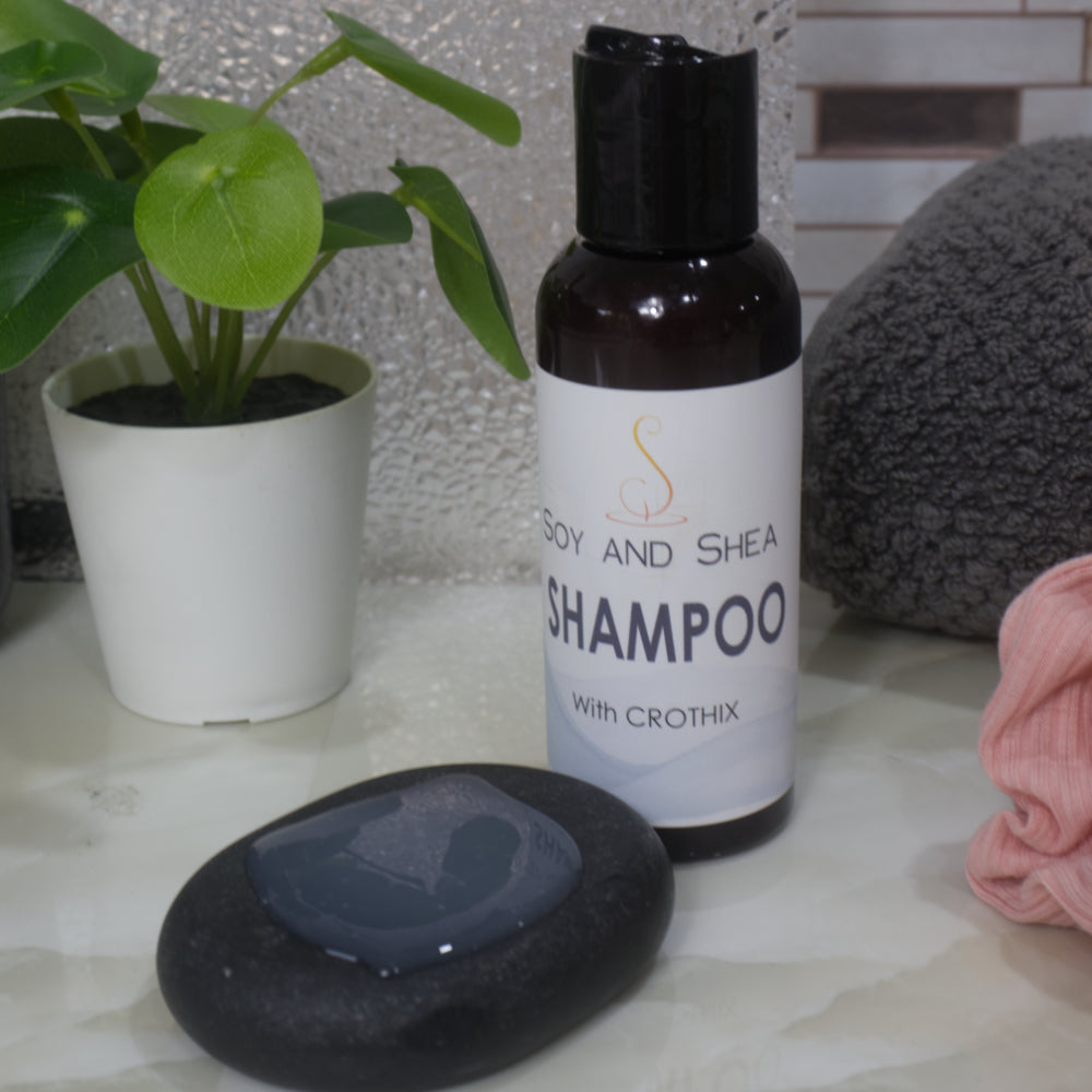 An amber bottle with disc cap lid stands on a bathroom counter top surrounded by a plant and towels.  The bottle is labelled as SHAMPOO.  To the front is a small black massage stone with some of the shampoo on it which is white with a translucent look.