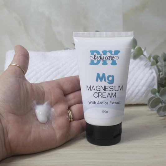 A white tube with black cap stands on a wooden surface with a towel and greenery in the background.  An open hand is next to the tube showing a small amount of white cream in the palm.