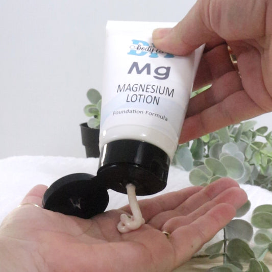 A hand squeezes a white tube with black flip top lid labelled as magnesium lotion into the palm of another hand.   The cream coming out of the tube has an off-white appearance.  The background is white with a green sprawling plant.