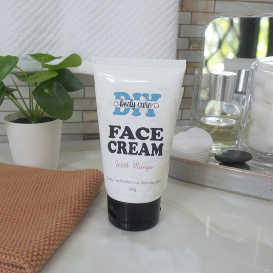 A white squeeze tube of DIY Body Care Face Cream with Mango sits on a white marble countertop in a bathroom setting, surrounded by a potted plant, a brown woven cloth, and skincare items in a mirror.