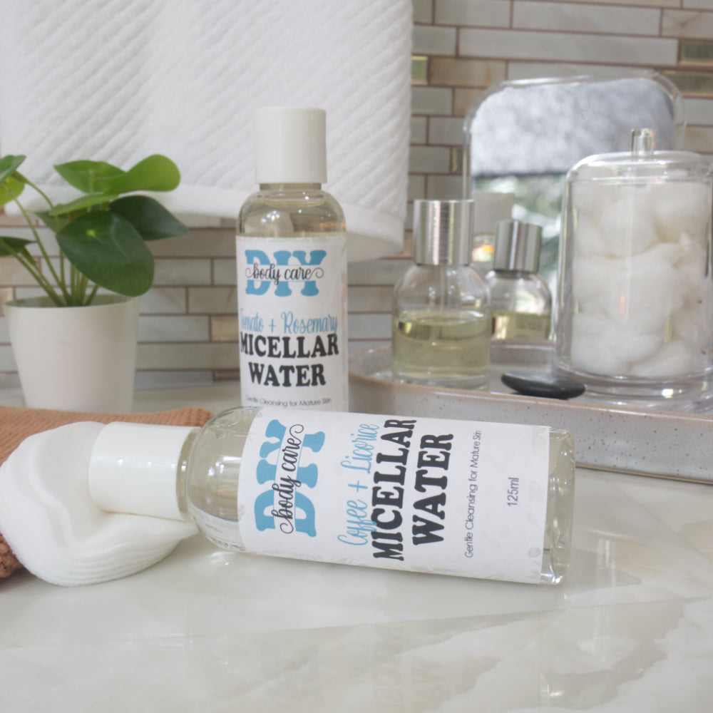 Two clear bottles with white caps are displayed on a bathroom counter top.  The bottle to the front lays on it side resting on a stack of cotton pads and is labelled as Coffee & Licorice Micellar Water.  Behind stands another bottle next to a plant and bottle of perfume.  It is labelled as Tomato & Rosemary Micellar Water
