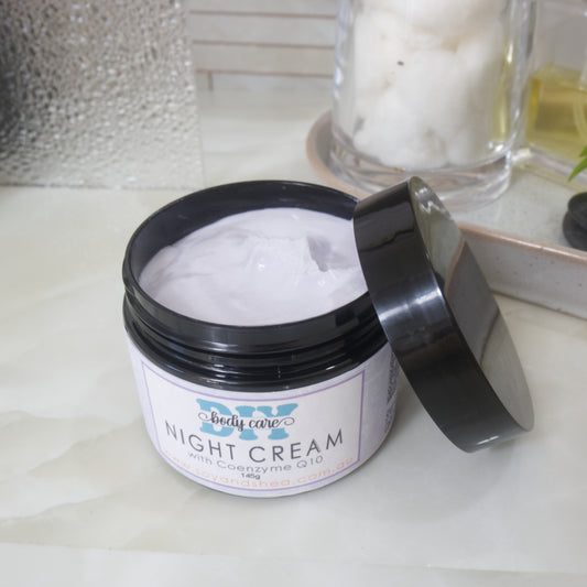 A black jar with a decorative purple label marked as NIGHT CREAM, sits on a bathroom counter top.  The lid has been removed and is resting on the side of the jar revealing a light purple cream inside.