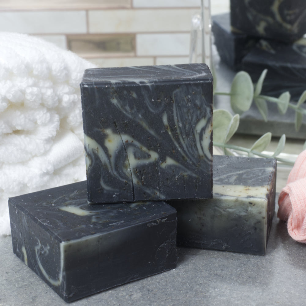 Two black square soaps with pale green swirls lay flat on a grey stone tray with a third soap standing upright upon them.   In the background is a white towel and a small mirror reflecting the soaps