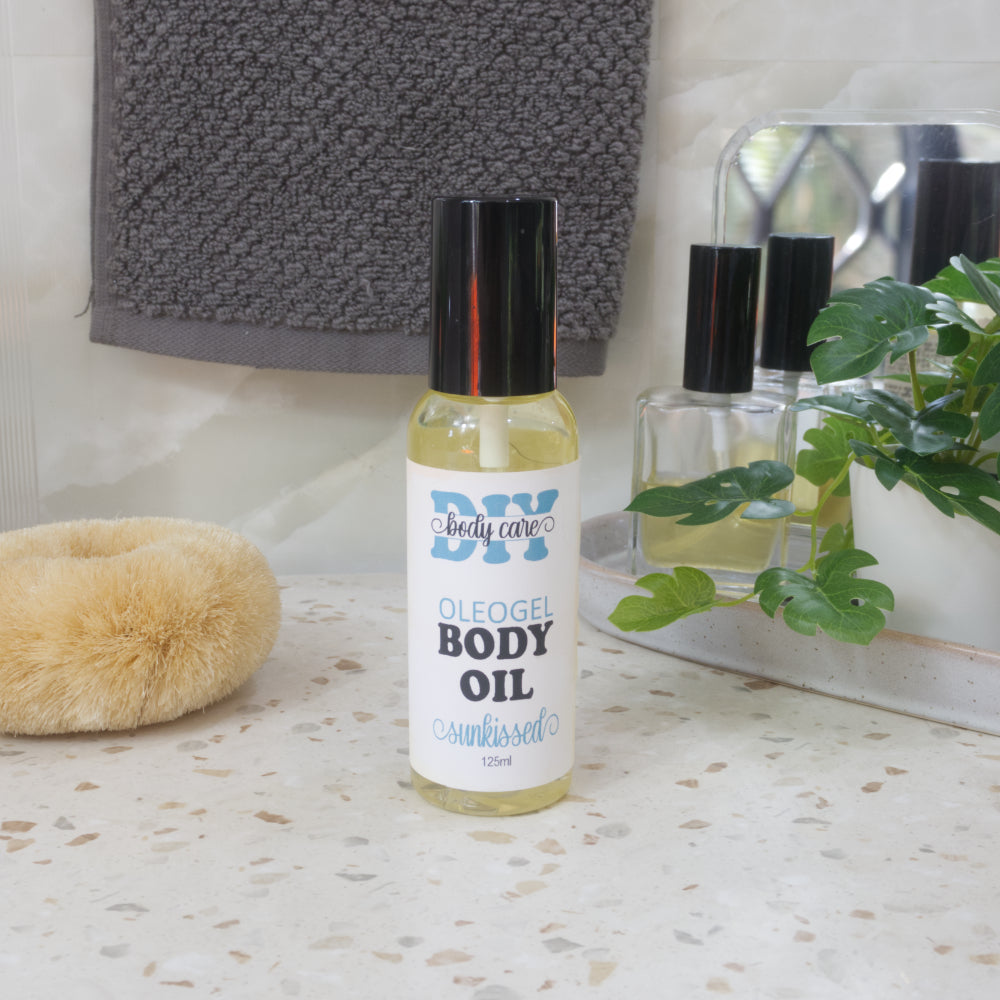 A clear bottle with a black lid sits on a bathroom counter top.  It is filled with a golden liquid and it has a product label identifying it as an Oleogel Body Oil in Sunkissed scent