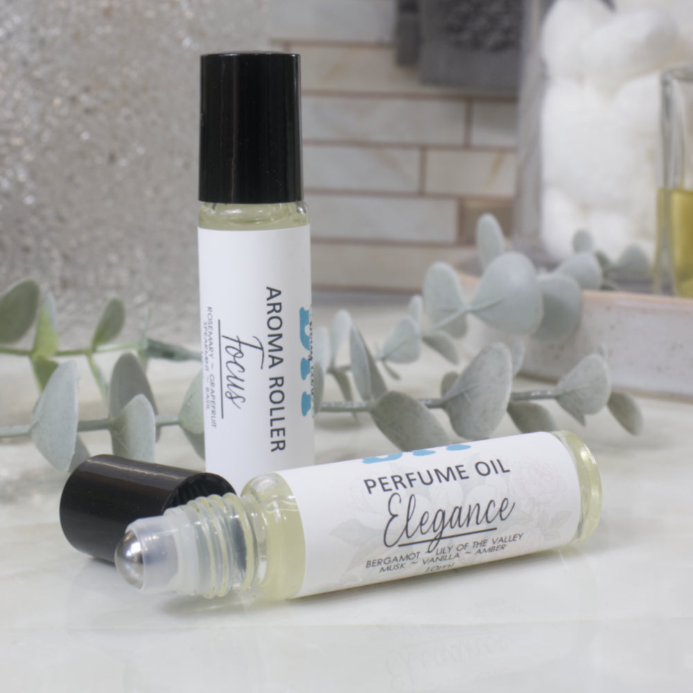 Two roll-on perfume oil bottles displayed on a light marble surface with eucalyptus leaves. One bottle stands upright with its black cap on, showing the label 'AROMA ROLLER Focus', and the other lies on its side showing 'PERFUME OIL Elegance'.  The black cap has been removed showign a metal ball roller applicator