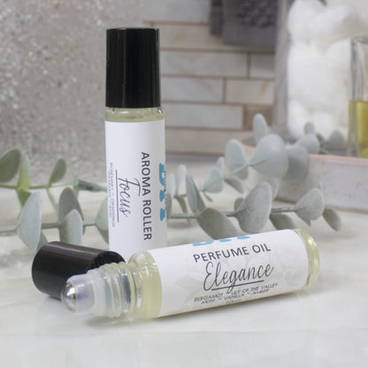 Two roll-on perfume oil bottles displayed on a light marble surface with eucalyptus leaves. One bottle stands upright with its black cap on, showing the label 'AROMA ROLLER Focus', and the other lies on its side showing 'PERFUME OIL Elegance'.  The black cap has been removed showign a metal ball roller applicator