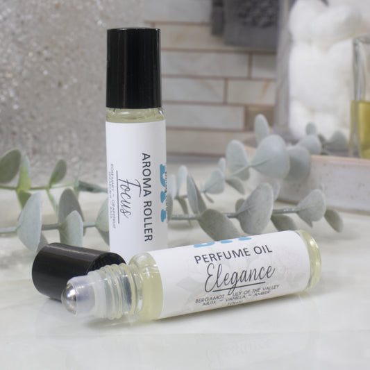 Two roll-on perfume oil bottles displayed on a light marble surface with eucalyptus leaves. One bottle stands upright with its black cap on, showing the label 'AROMA ROLLER Focus', and the other lies on its side showing 'PERFUME OIL Elegance'.  The black cap has been removed showign a metal ball roller applicator