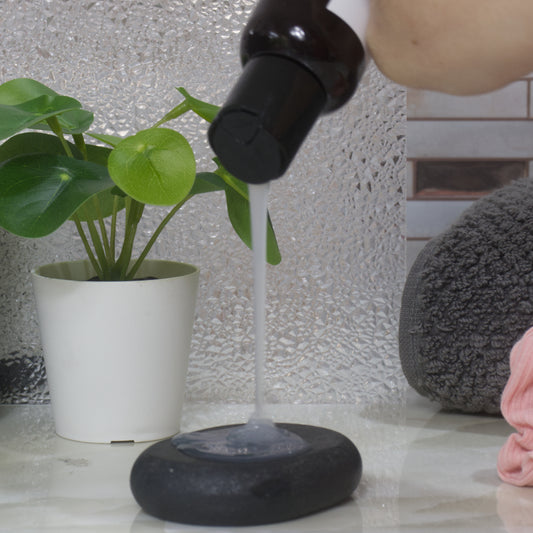 Person pouring a white translucent liquid from a bottle onto a black stone that rests on a marble surface with a plant and towel in the background.