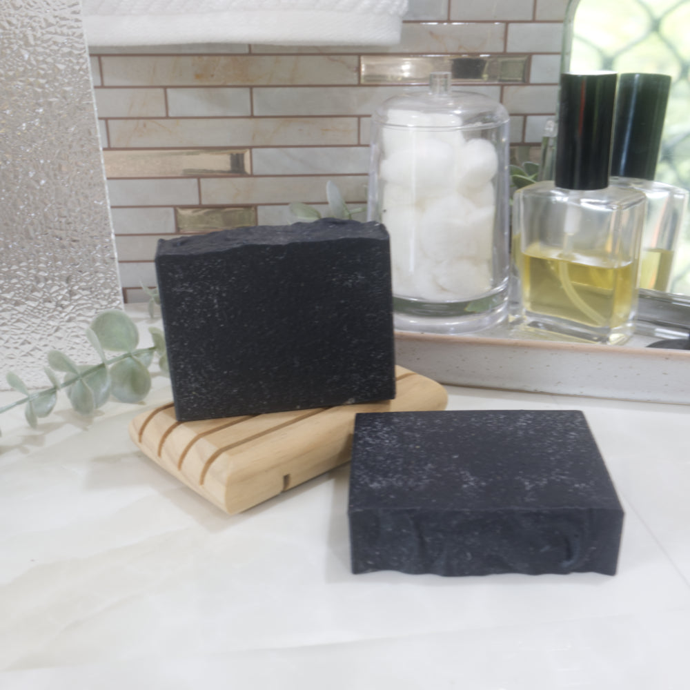 A black soap bar stands on a wooden soap rack with another laying flay on a marble surface just in front of the soap rack.  Behind the soaps are bathroom items like a jar of cotton balls, and perfume bottle.