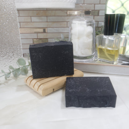 A black soap bar stands on a wooden soap rack with another laying flay on a marble surface just in front of the soap rack.  Behind the soaps are bathroom items like a jar of cotton balls, and perfume bottle.