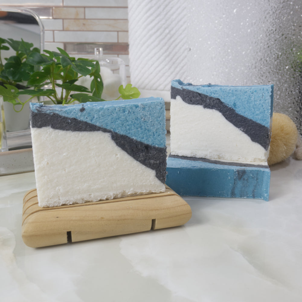 Three soaps are on a bathroom countertop.  One soap lays flat on the bench showing a flat teal top.  Another soap stands up on this soap and the left is another soap on a wooden soap dish.  The soaps have a diagonal strip pattern with a large white area witha thin layer of black then finished with teal
