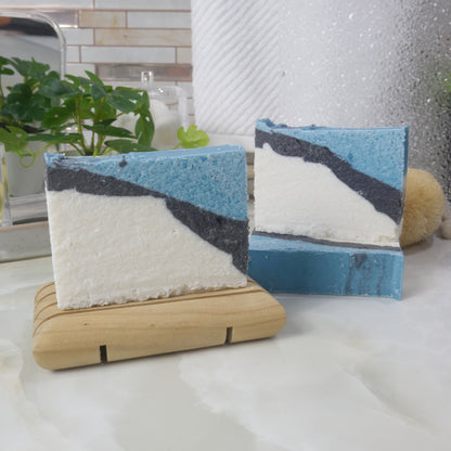 Three soaps are on a bathroom countertop.  One soap lays flat on the bench showing a flat teal top.  Another soap stands up on this soap and the left is another soap on a wooden soap dish.  The soaps have a diagonal strip pattern with a large white area witha thin layer of black then finished with teal