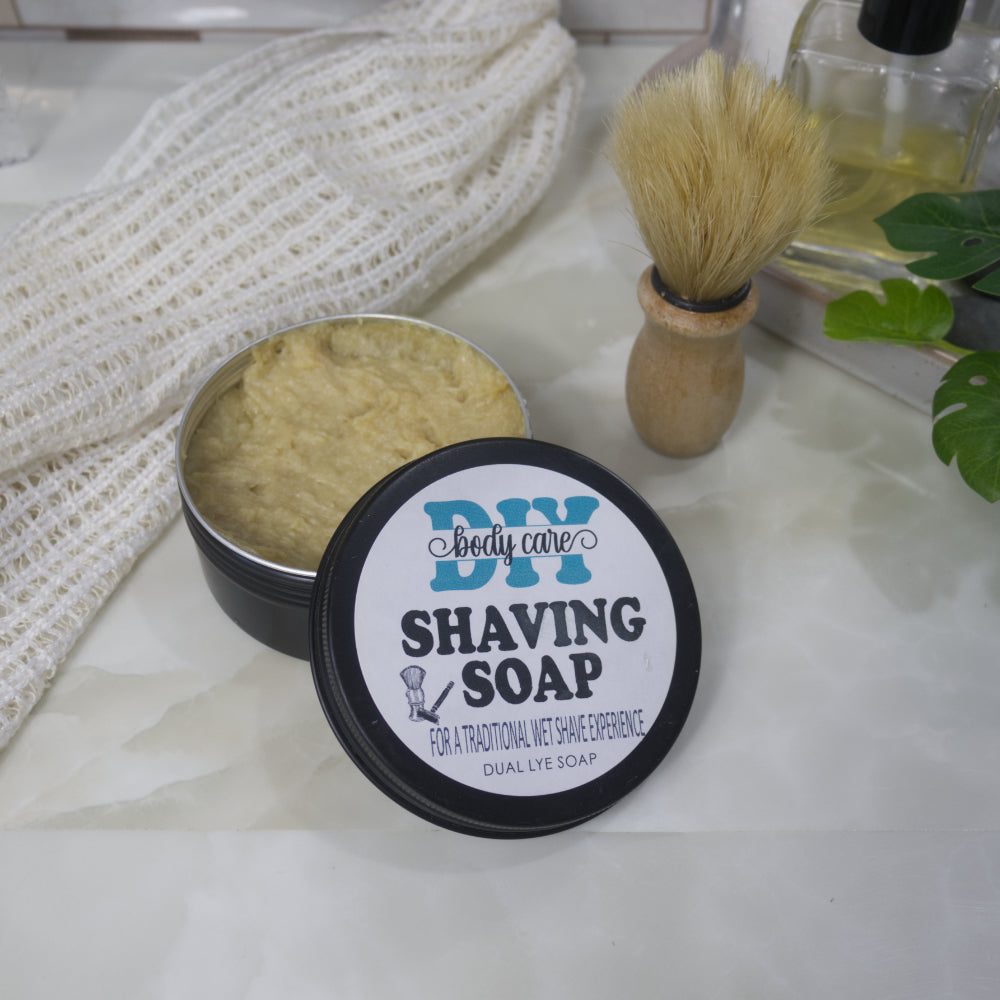 A black round tin sits on a bathroom counter top next to a shaving brush and cotton cloth.  The lid, which is labelled as Shaving Soap, has been removed and rests on the side of the tin.  The tin is filled with a beige coloured soap.