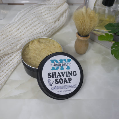A black round tin sits on a bathroom counter top next to a shaving brush and cotton cloth.  The lid, which is labelled as Shaving Soap, has been removed and rests on the side of the tin.  The tin is filled with a beige coloured soap.