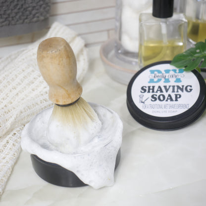 A round tin sits on a bathroom counter top with cotton cloth.  The tin has a dense, white lather on it witha shaving brush standing in the middle with bristles down in the foam.   To the right i the lid with a label identifying it as Shaving Soap