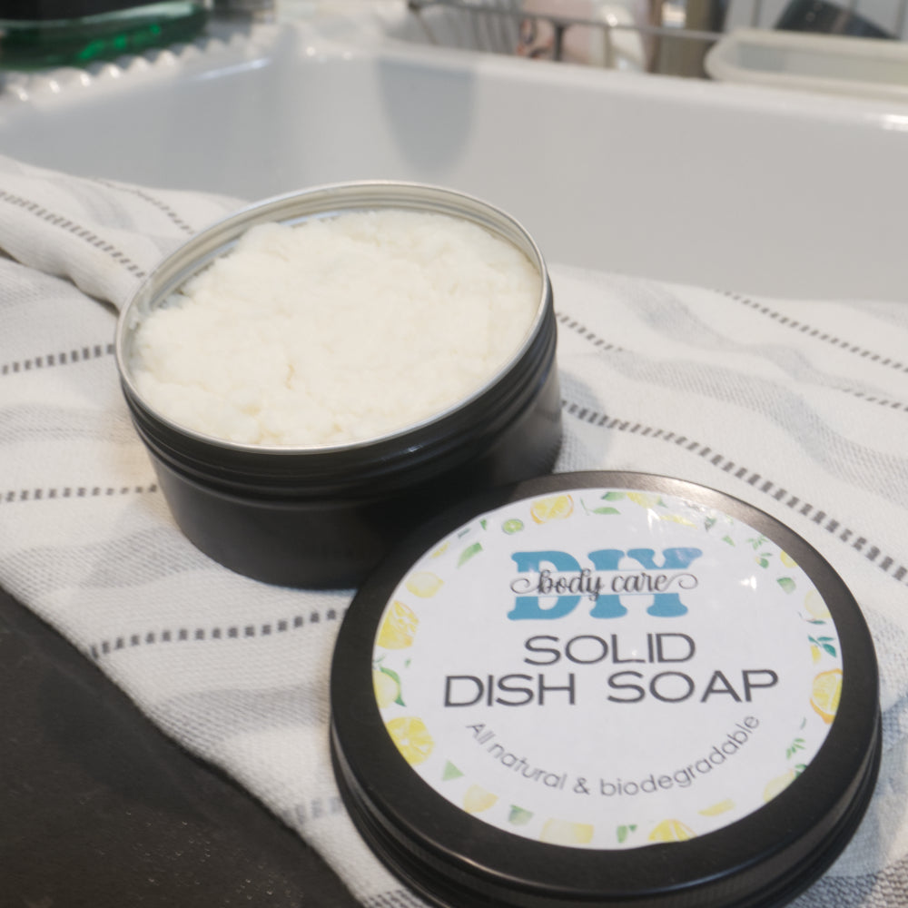 A round black tin sits on a dish towel next to a white kitchen sink with a blurred background of a dish rack.  The lid of the has been removed to reveal a white paste.  The lid sits next to the tin with a product label identifying it as Solid Dish Soap