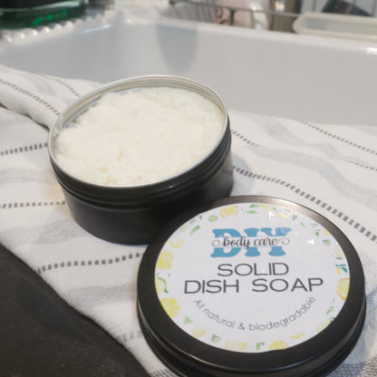 A round black tin sits on a dish towel next to a white kitchen sink with a blurred background of a dish rack.  The lid of the has been removed to reveal a white paste.  The lid sits next to the tin with a product label identifying it as Solid Dish Soap