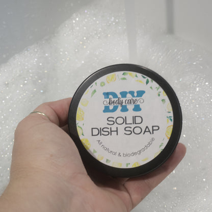 A hand holds a round black tin with a label reading Solid Dish Soap by DIY Body Care.  Behind is a white sink with water flowing into it and a mound of bubbles.