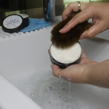A person holds an open tin of solid dish soap over a white sink.  They use a scrubbing brush in the other hand to build up a lather.