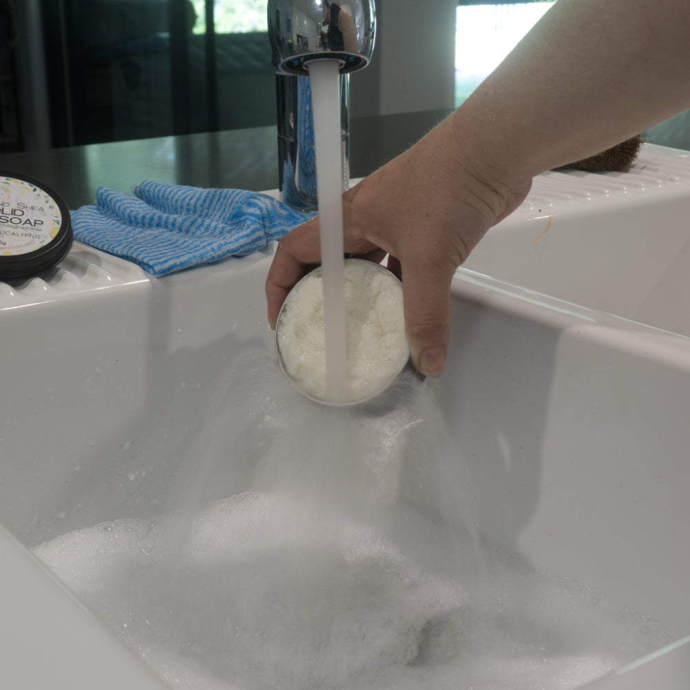 A hand holds a tin of white solid dish soap under a tap of running water.   The water is running the tin into a white sink creating a mass of bubbles.