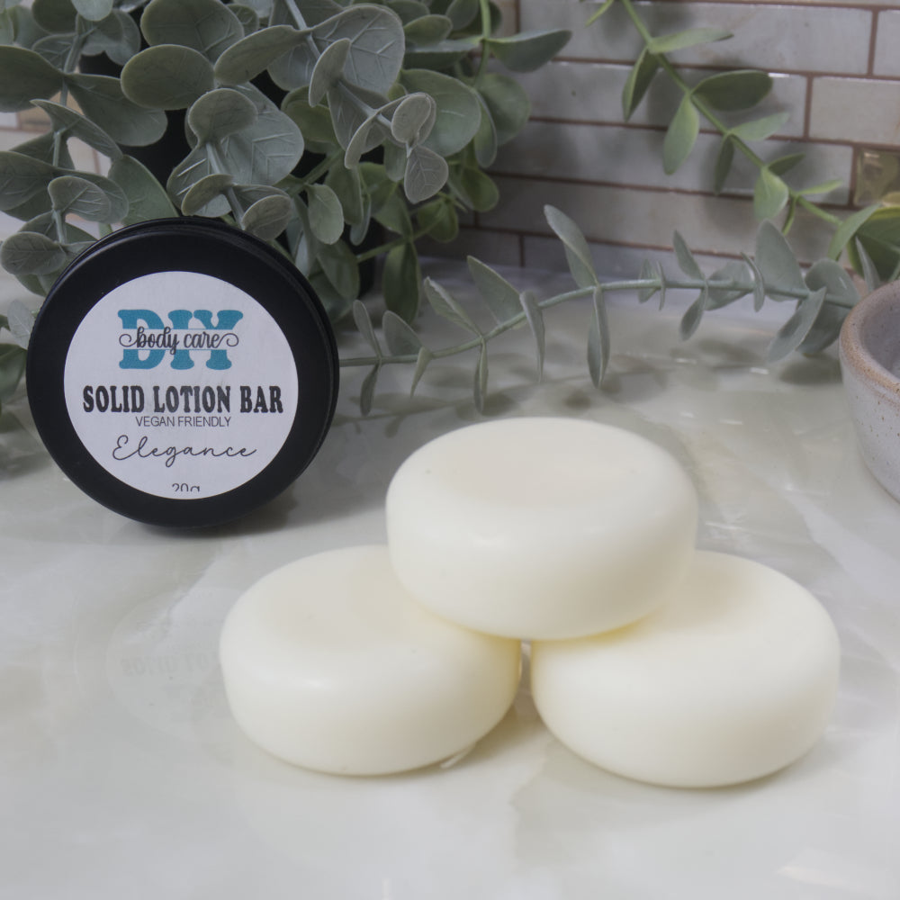 A pyramid stack of three white round pucks sit in the foreground on a marble surface.  Behind is a black tin standing on its side with a label reading Solid Lotion Bar.  It is surrounded by greenery and a tile background.