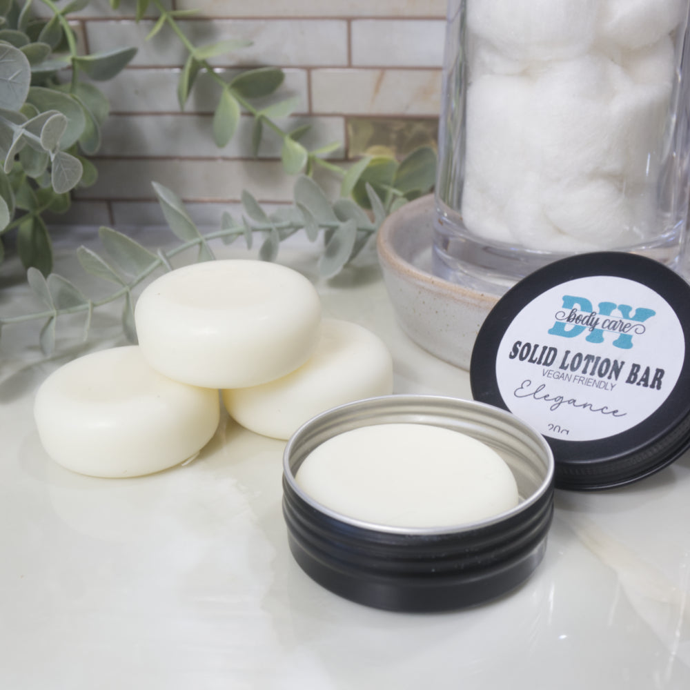 A round black tin sits on a marble surface with its lid resting against a stone tray.  Inside the tin is a white puck and the lid is labelled as Solid Lotion Bar.  Behind the tin is a pyramid stack of three more white pucks surrounded by greenery