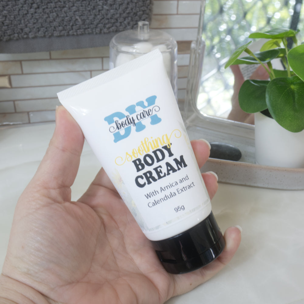 A hand holding a white squeeze tube of DIY Body Care Soothing Body Cream with a black cap, set against a softly blurred bathroom background with a plant and mirror.