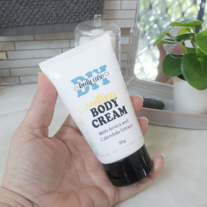 A hand holding a white squeeze tube of DIY Body Care Soothing Body Cream with a black cap, set against a softly blurred bathroom background with a plant and mirror.