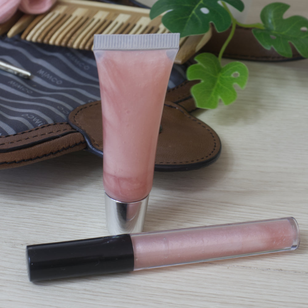 Two lip oils on a wooden surface with a plant and open handbag in the background.  One lip oil is in a cylindrical tube with black lid while the other is in a small tube with silver lid.  Both are clear packaging showing a pale pink liquid inside.