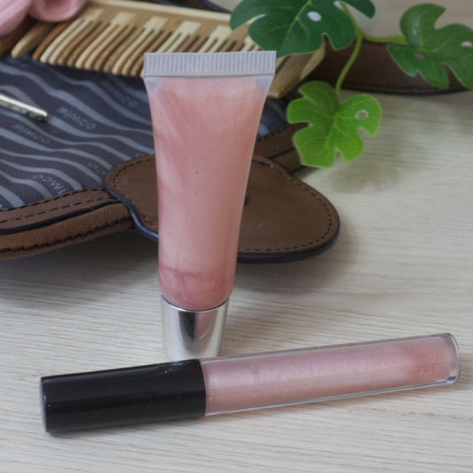 Two lip oils on a wooden surface with a plant and open handbag in the background.  One lip oil is in a cylindrical tube with black lid while the other is in a small tube with silver lid.  Both are clear packaging showing a pale pink liquid inside.