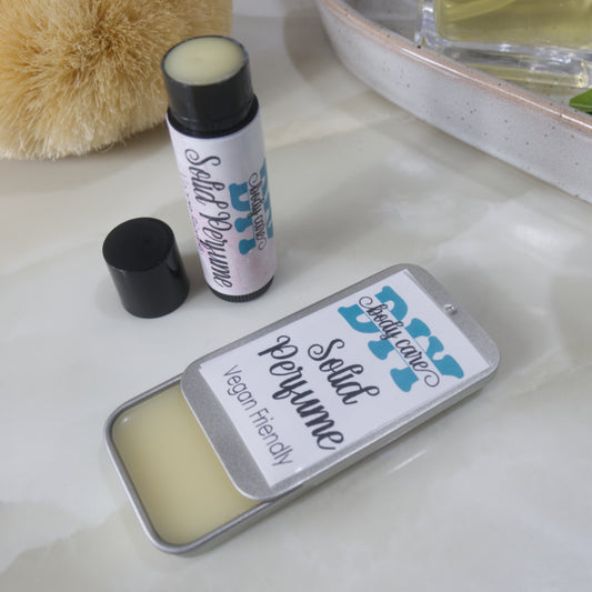 Two solid perfume are sitting on a bathroom countertop.  One is presented in a rectangular silver tin with a open sliding lid while the other is a black tube like lip balm, standing upright with the lid to the side of the tube revealing white balm inside.