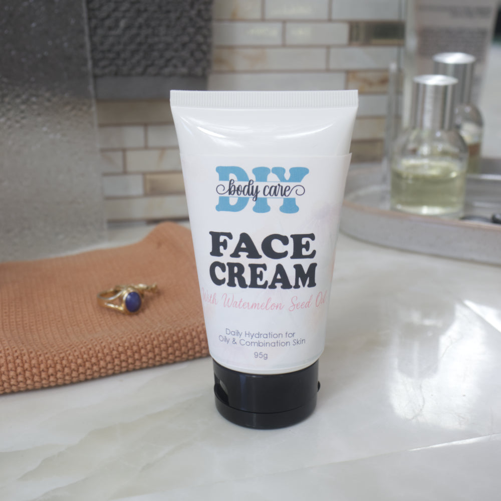 A white squeeze tube of DIY Body Care Face Cream with Watermelon Seed Oil, featuring a black cap, standing on a white marble countertop with a textured orange towel and bathroom background.