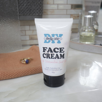 A white squeeze tube of DIY Body Care Face Cream with Watermelon Seed Oil, featuring a black cap, standing on a white marble countertop with a textured orange towel and bathroom background.
